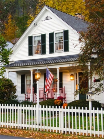 Two story country home with white picket fence, American flag and red rocking chairs.